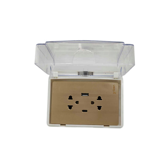 A gold-plated wall outlet socket with a built-in USB port, enclosed in a clear plastic protective cover that is shown open, on a plain white background.