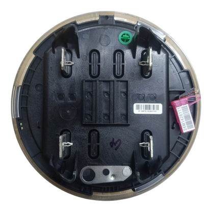The underside of a circular black plastic device, likely a smart meter or utility reader, featuring metal terminal contacts, a barcode label, and a small green screw. A pink and white tag is attached on the right side, all set against a plain white background.