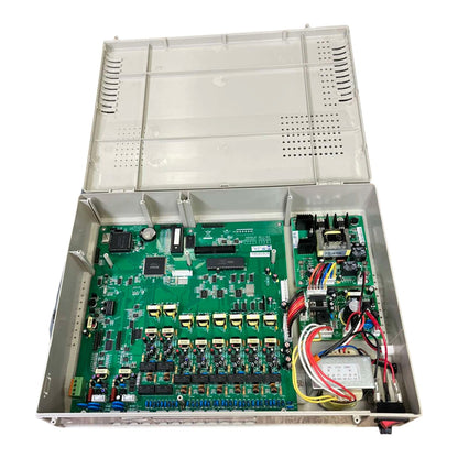 Open industrial electronic control cabinet showing a large green circuit board with multiple modules, connectors, and wiring harnesses inside a metal housing, photographed on a white background.
