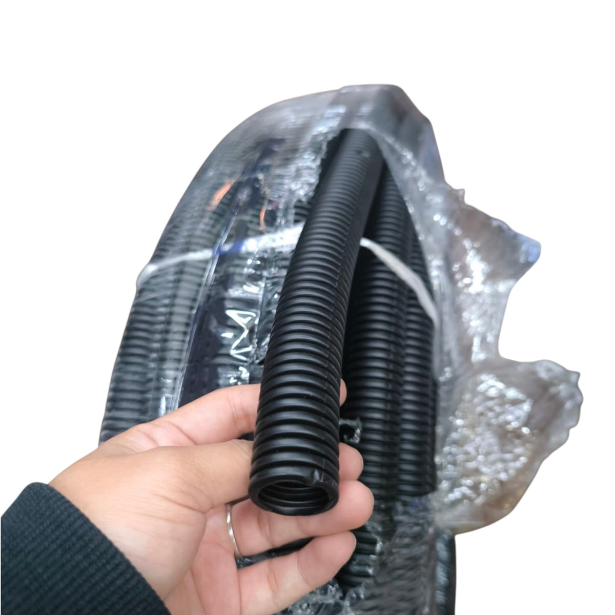 A hand holding a short section of black corrugated plastic conduit, with a larger spool of the same black tubing wrapped in clear protective plastic behind it on a white background.