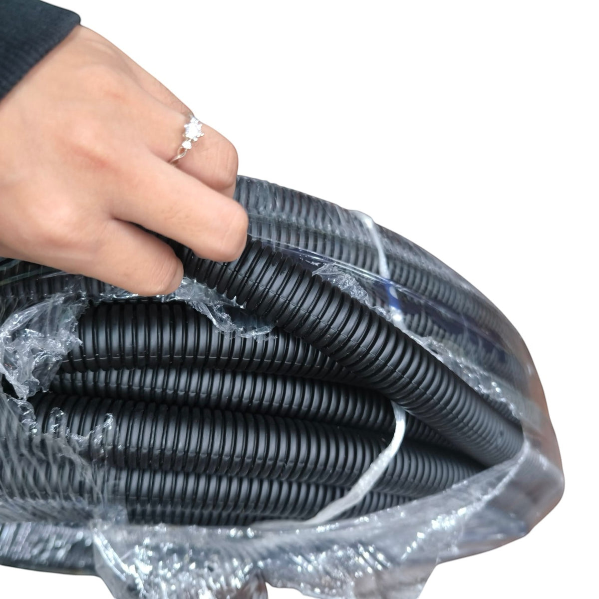 A close-up of a person's hand holding a coiled black corrugated plastic conduit or cable protective tube wrapped in clear plastic, against a plain white background.