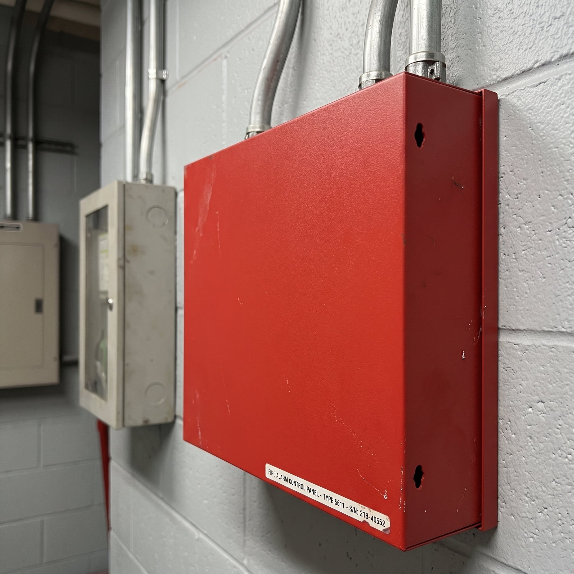 A red metal electrical junction box mounted on a light gray concrete block wall, with metal conduit pipes entering from the top and a small white label near the bottom edge.