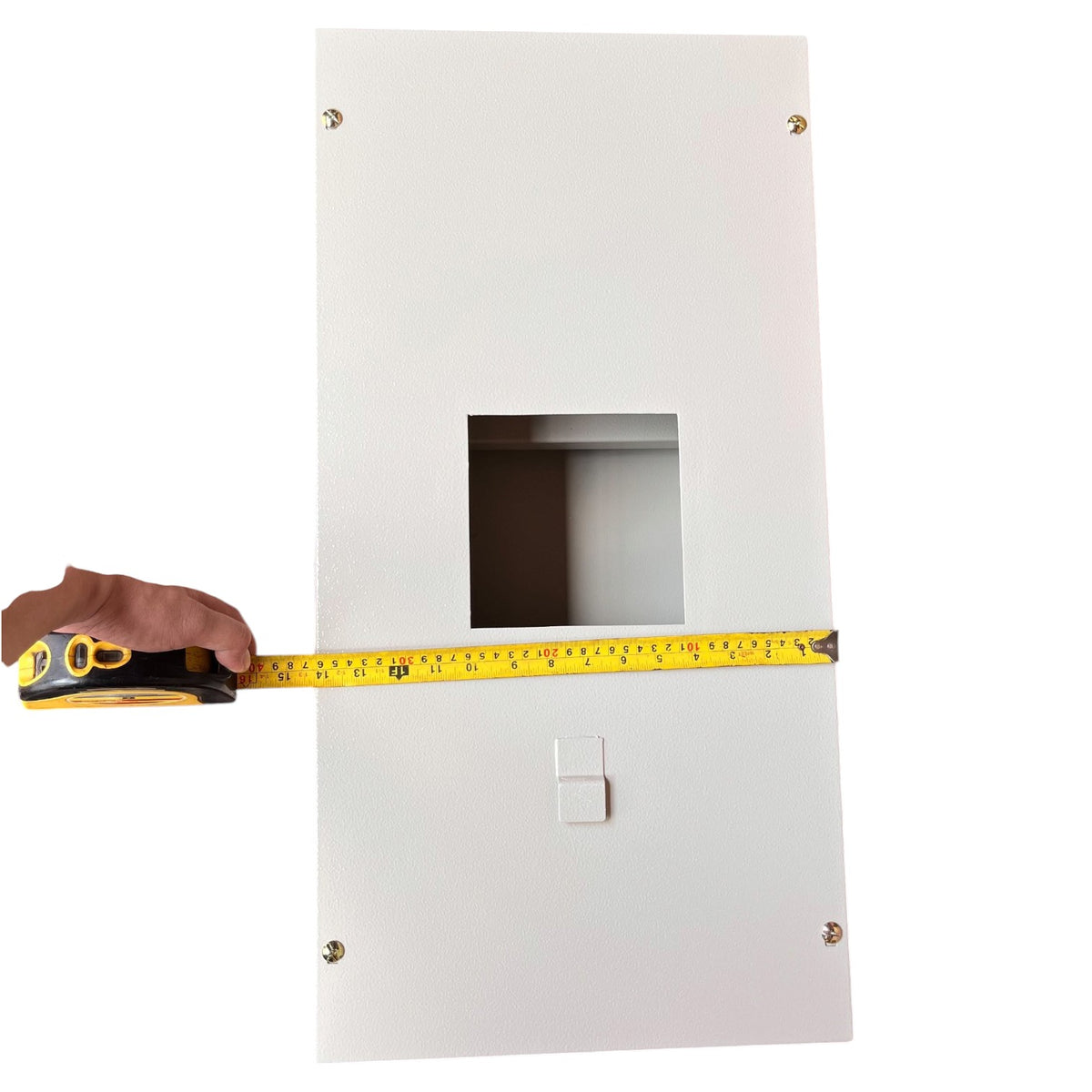 A white rectangular electrical or utility panel with a square cutout in the center and a small latch near the bottom, shown with a hand holding a yellow tape measure across it to indicate width.
