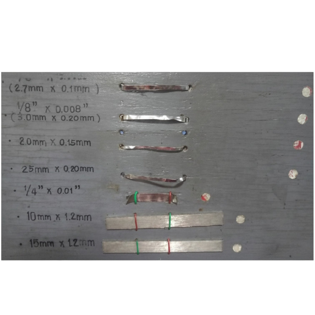 A gray metal sample board with handwritten size measurements in millimeters and inches next to several small strips of metallic material, likely showing material samples by size.