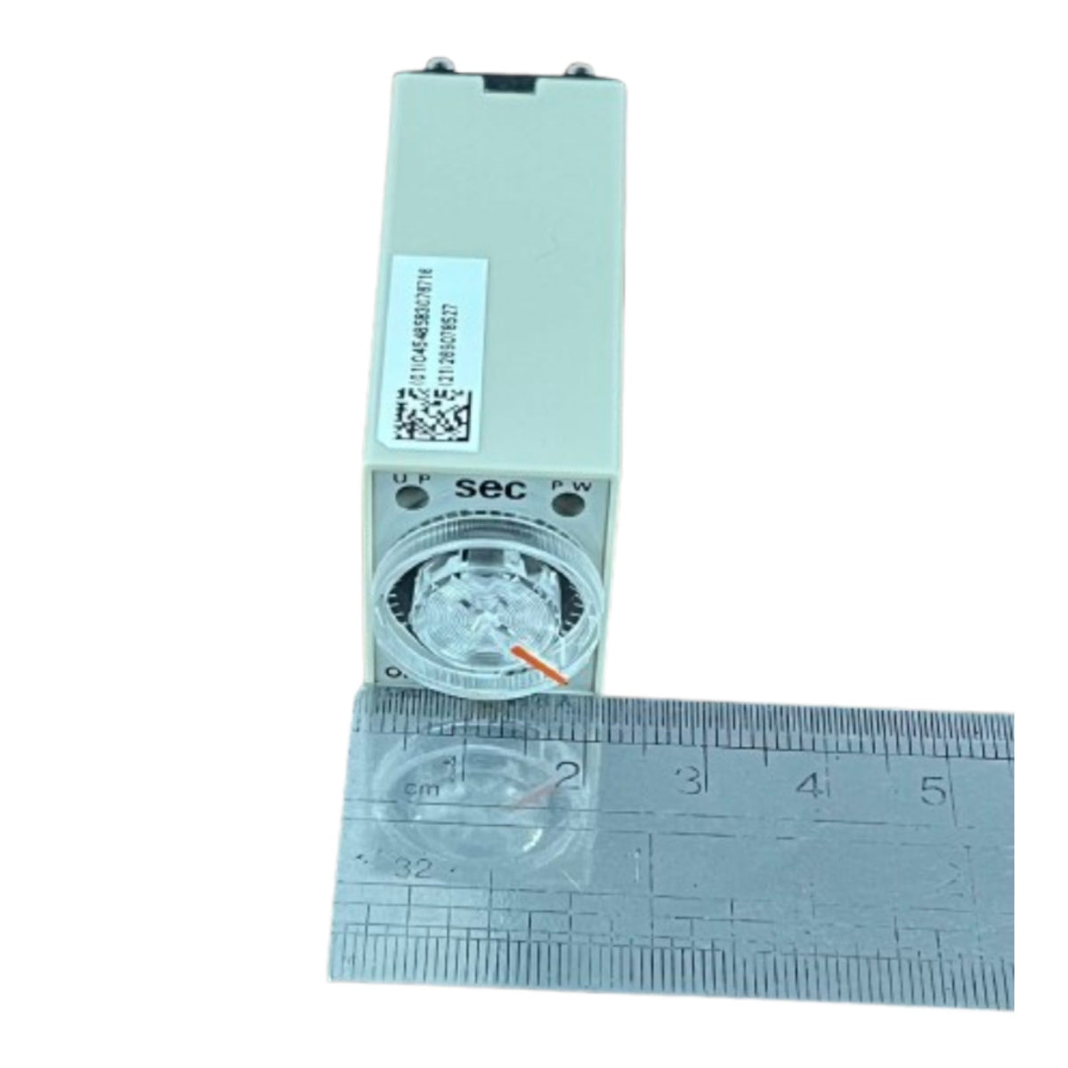 A small rectangular electronic device with a circular dial or window on the front, shown upright next to a metal ruler for scale on a plain white background.