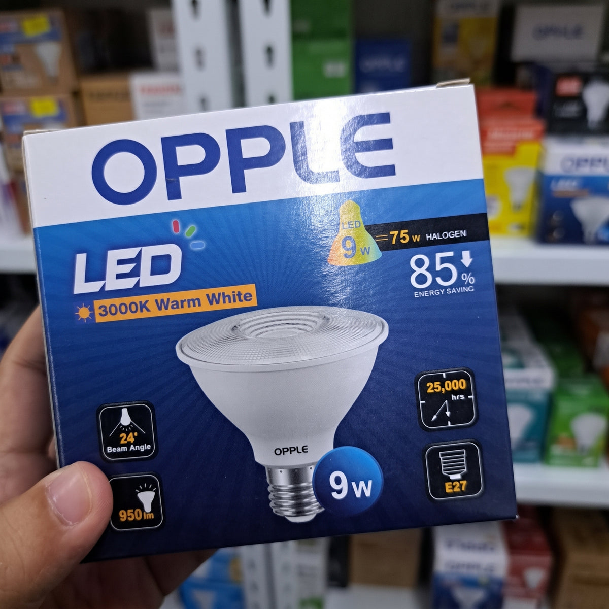 A hand holding an OPPLE LED light bulb box in a retail store. The box is blue and white, featuring a picture of a 9-watt warm white LED bulb and product specifications.