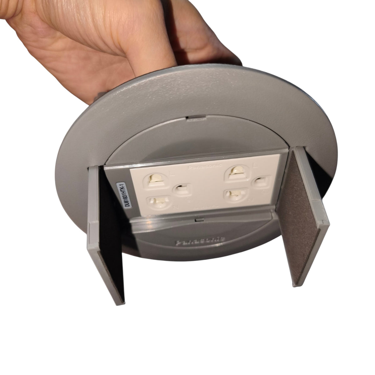 A hand holding a round gray recessed outlet cover with two flaps open, revealing a white four-outlet electrical outlet underneath, on a plain white background.