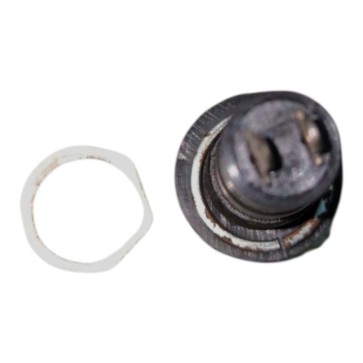 A small circular metal component with a dark, worn surface and a separate white ring, shown on a plain white background.