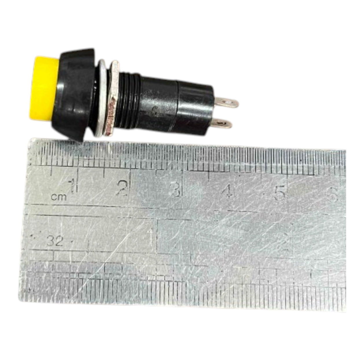 A small black electrical push-button switch with a yellow top and two metal pins, shown next to a metal ruler for scale on a white background.