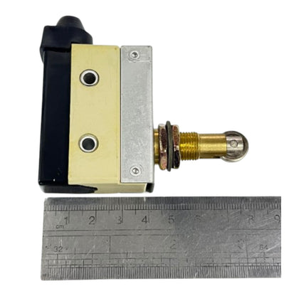 A small mechanical switch or valve component with a black and brass-colored body, shown next to a metal ruler for scale on a white background.
