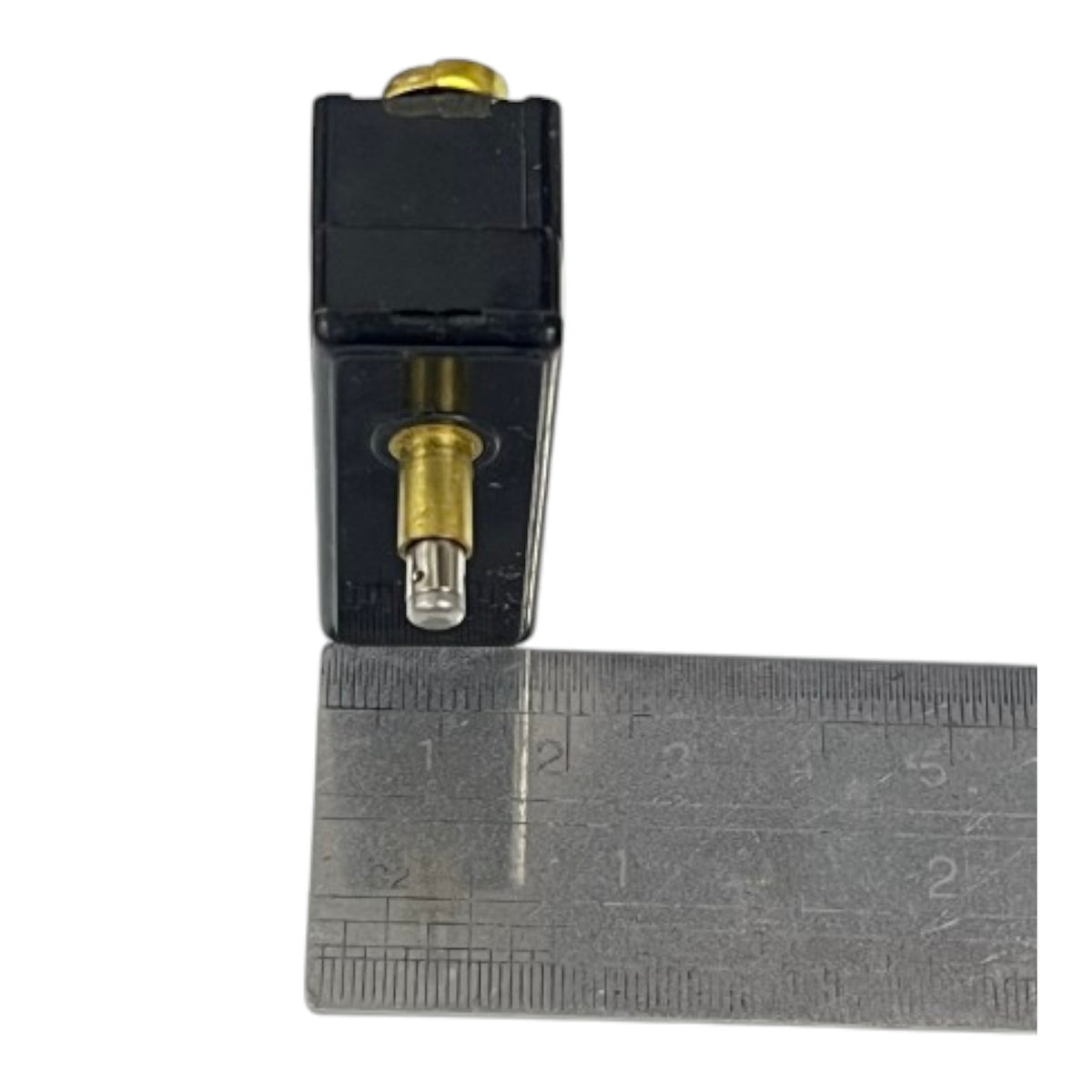 A small black electronic component with brass-colored metal contacts and a clear lens, shown next to a metal ruler for scale on a white background.